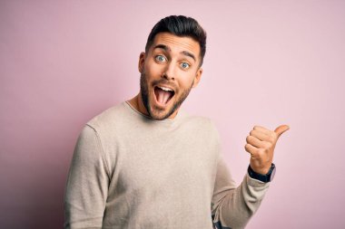 Soyutlanmış pembe arka planda dikilen, mutlu yüzüyle gülümseyen ve baş parmağıyla yan tarafı gösteren sıradan bir süveter giyen genç yakışıklı adam..