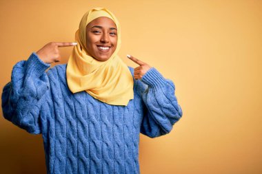 Genç Afrikalı Afro kadın, izole edilmiş sarı arka planda Müslüman başörtüsü takıyor neşeli gülümsüyor ve parmaklarını ve ağzını işaret ediyor. Diş sağlığı konsepti.