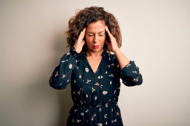 Orta yaşlı, güzel bir kadın, izole edilmiş beyaz arka planda dikilip acı ve migren yüzünden çaresiz ve stresli bir şekilde acı çeken sıradan bir elbise giyiyor. Ellerini başının üstüne koy..