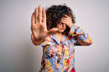 Genç ve güzel kıvırcık Arap kadın çiçek rengi gömlek giyiyor beyaz arka planda duruyor gözleri ellerle örtüyor ve üzgün ve korku ifadeleriyle dur hareketi yapıyor. Utanç verici ve olumsuz bir kavram..