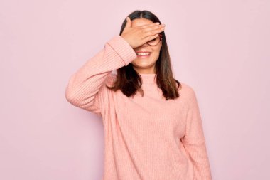 Genç, güzel, esmer bir kadın pembe arka planda gözlük ve süveter takıyor, gülümsüyor ve yüzünde şaşkınlıktan gözleri kapayarak gülüyor. Kör kavram.