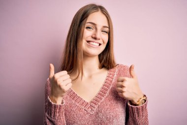 Genç, güzel, kızıl saçlı bir kadın pembe arka plan başarısı tabelasının üzerine günlük süveter giyiyor. Elleriyle olumlu jest yapıyor, gülümsüyor ve mutlu. Neşeli ifade ve kazanan jest.