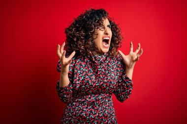 Genç, güzel, kıvırcık, çiçekli bir elbise giymiş, kırmızı arka planda dikilmiş deli deli deli bağırıyor ve agresif bir ifade ve kolla bağırıyor. Hayal kırıklığı konsepti.
