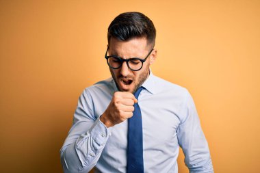 Sarı arka plan üzerinde duran kravat ve gözlük takan genç yakışıklı işadamı kendini iyi hissetmiyor ve soğuk algınlığı veya bronşitin belirtisi olarak öksürüyor. Sağlık hizmeti kavramı.