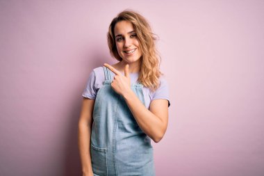Genç, güzel, sarışın bir kadın pembe arka planda dikilen rahat bir kot pantolon giyiyor. Yüzünde mutlu ve doğal bir ifadeyle gülümseyen bir yüz ifadesi var.