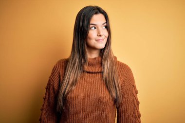Genç, güzel bir kız, günlük süveter giyer, izole edilmiş sarı arka planda durur, gülümser ve başka tarafa bakar ve düşünür..