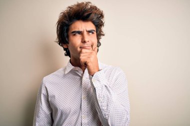 Zarif bir gömlek giyen yakışıklı bir iş adamı, soyutlanmış beyaz arka planda, elleri çenesinde soru sorarken, düşünceli bir ifade takınıyor. Düşünceli bir yüzle gülümsüyor. Şüphe kavramı.