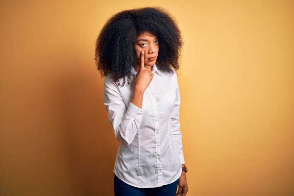 Young beautiful african american elegant woman with afro hair standing over yellow background Pointing to the eye watching you gesture, suspicious expression