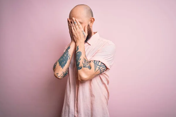 Handsome bald man with beard and tattoo wearing casual shirt over isolated pink background with sad expression covering face with hands while crying. Depression concept.