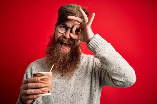 Irish redhead man with beard drinking a takeaway coffee on a paper cup over red background with happy face smiling doing ok sign with hand on eye looking through fingers