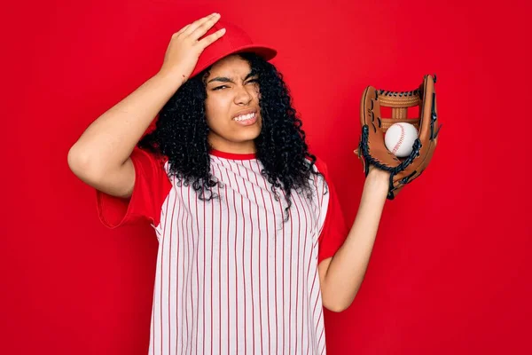 Young african american curly sportswoman wearing cap playing baseball using ball and glove stressed with hand on head, shocked with shame and surprise face, angry and frustrated. Fear and upset for mistake.