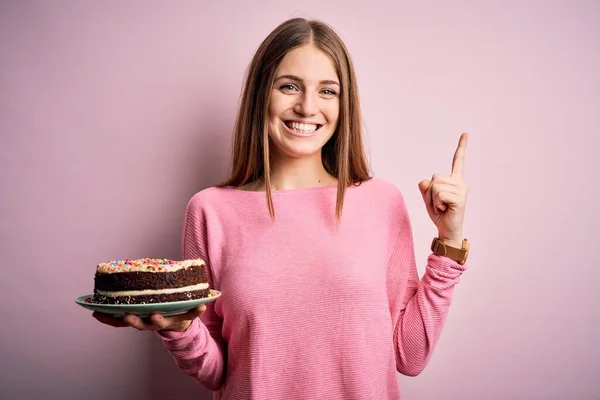 若いです美しいです赤毛女性保持誕生日ケーキ以上隔離されたピンクの背景驚きでAアイデアや質問指差し指で幸せな顔 ナンバーワン — ストック写真