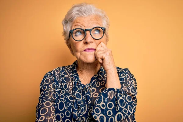 Senior beautiful grey-haired woman wearing casual shirt and glasses over yellow background with hand on chin thinking about question, pensive expression. Smiling with thoughtful face. Doubt concept.