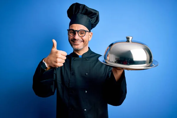 年轻英俊的厨师 身穿制服 头戴礼帽 拿着餐盘 顶着圆顶 喜形于色 笑容满面 做手势 用手指竖起大拇指 很好的标志 — 图库照片