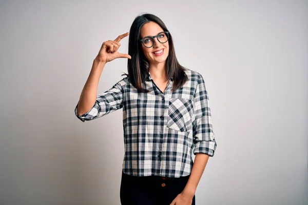 Young brunette woman with blue eyes wearing casual shirt and glasses over white background smiling and confident gesturing with hand doing small size sign with fingers looking and the camera. Measure concept.