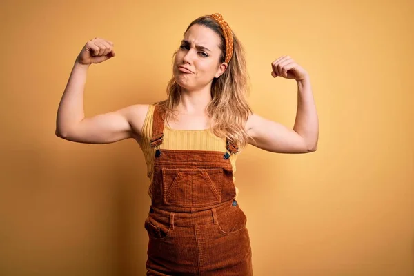 Young beautiful blonde woman wearing overalls and diadem standing over yellow background showing arms muscles smiling proud. Fitness concept.