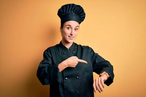 年轻美丽的厨师女士身穿炊具制服 头戴黄底帽 匆忙地指着时间 不耐烦 烦躁不安 对最后期限的延误感到愤怒 — 图库照片