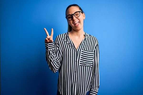 Beautiful blonde woman with blue eyes wearing striped shirt and glasses over blue background smiling with happy face winking at the camera doing victory sign. Number two.