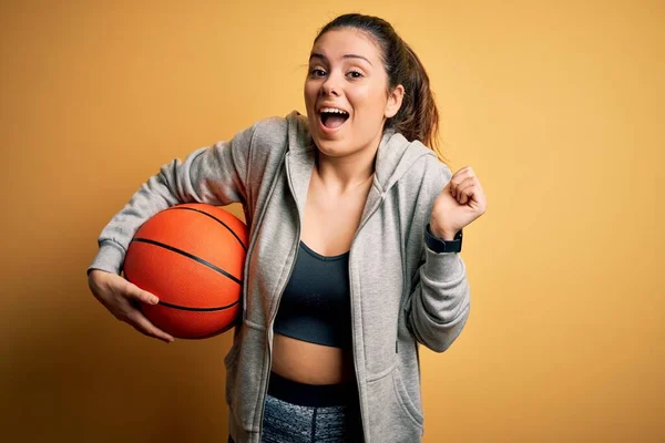 年轻美丽的黑发女运动员在黄色的背景下举行篮球比赛 她自豪地尖叫着 为胜利和成功欢呼着 激动地欢呼着 — 图库照片