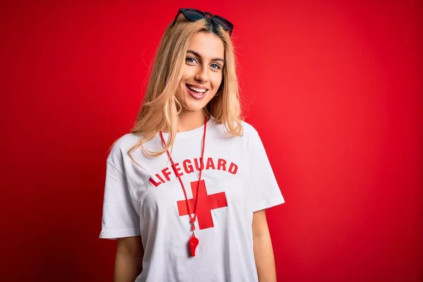 Young beautiful blonde lifeguard woman wearing t-shirt with red cross using whistle with a happy and cool smile on face. Lucky person.