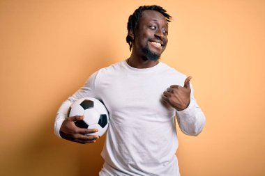 Genç Afrikalı Amerikalı oyuncu futbol topunu sarı arka planda tutuyor ve gülümseyen bir gülümsemeyle baş parmağıyla işaret ediyor.