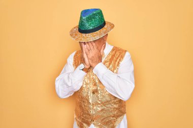 Sarı arka planda Brezilya karnavalı kıyafeti giyen orta yaşlı, kır saçlı bir adam. Ağlarken suratını ellerinle kapatan üzgün bir ifade. Depresyon kavramı.