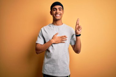 Yakışıklı Afro-Amerikan sporcusu spor giyer sarı arka planda spor giyer gülümser, el ve göğsü havada küfreder, sadakat yemini eder.