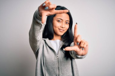 Genç, güzel Çinli sporcu kadın izole edilmiş beyaz arka plan üzerine kazak giyip gülümsüyor ve elleri ve parmakları gülen yüzlü bir çerçeve yapıyor. Yaratıcılık ve fotoğrafçılık kavramı.