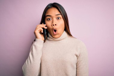 Genç Asyalı kız akıllı telefondan konuşuyor pembe arka planda, şok olmuş, korkmuş ve korku ifadesinden heyecanlanmış bir şekilde.