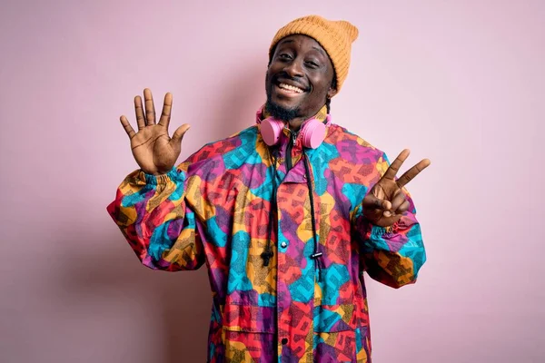 Young handsome african american man wearing colorful coat and cap over pink background showing and pointing up with fingers number seven while smiling confident and happy.