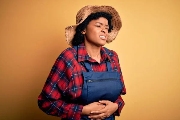 Young African American afro farmer woman with curly hair wearing apron and hat with hand on stomach because nausea, painful disease feeling unwell. Ache concept.