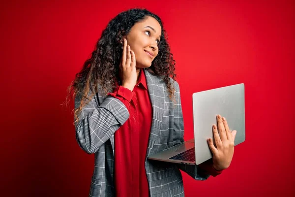 Young beautiful business woman with curly hair working using laptop over red background serious face thinking about question, very confused idea