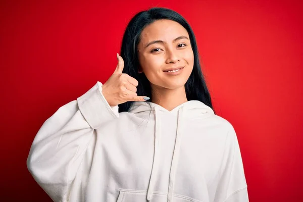 年轻美丽的中国女运动员穿着运动衫 站在孤立的红色背景上 微笑着 用手和手指做着电话手势 就像在讲电话一样 交流概念 — 图库照片