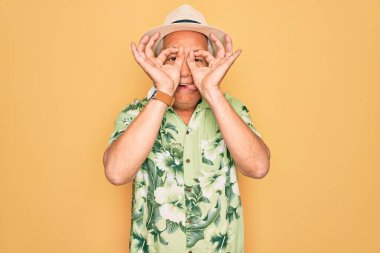 Orta yaşlı, kır saçlı, yaz tatilinde şapkalı ve çiçekli gömlek giyen dürbün gibi jest yapan, parmaklarından bakan yaşlı bir adam. Çılgın ifade.