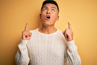 Sarı arka planda beyaz süveter giyen genç, yakışıklı Latin adam şaşırdı ve şaşırdı. Parmaklarıyla işaret etti ve kollarını kaldırdı..