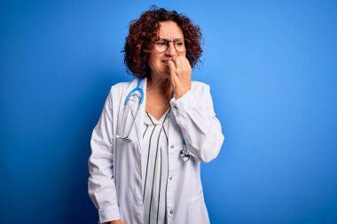 Mavi arka plan üzerinde steteskop ve ceket giyen orta yaşlı kıvırcık saçlı kadın elleri ağız yiyen tırnaklarla stresli ve gergin görünüyor. Endişe sorunu.