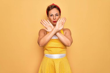 Orta yaşlı, son sınıf rozetli, 50 'li yaşlarda eski tarz elbise giyen, sarı arka planda reddedilmiş, kollarını kavuşturarak negatif işaret veren, kızgın yüzlü bir kadın.