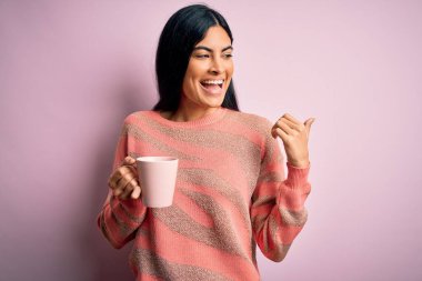Genç, güzel İspanyol bir kadın pembe izole arka planda bir fincan sıcak kahve içiyor ve gülümseyen bir gülümsemeyle baş parmağıyla onu işaret ediyor.