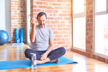 Orta yaşlı yakışıklı sporcu mindere oturmuş spor salonunda yoga egzersizi yapıyor. İtalyan jestleri yapıyor. Elleri ve parmakları kendinden emin ifade ediyor.