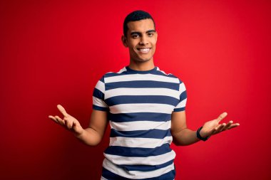 Yakışıklı afro-amerikan adam, günlük çizgili tişört giyiyor kırmızı arka planda gülümsüyor ve gülümsüyor, dostça karşılanıyor, olumlu ve kendinden emin selamlar.