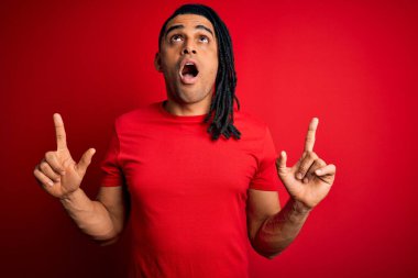 Kırmızı günlük tişörtlü Afro-Amerikan genç yakışıklı adam şaşkın şaşkın yukarı bakıyor ve parmaklarıyla işaret ediyor ve kollarını kaldırıyor..