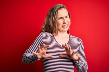 Young beautiful blonde woman wearing casual striped t-shirt over isolated red background disgusted expression, displeased and fearful doing disgust face because aversion reaction. With hands raised