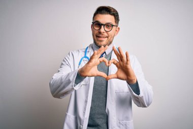 Mavi gözlü genç doktor tıbbi ceket ve steteskop takıp izole edilmiş arka planda gülümsüyor ve elleriyle kalp sembolü çiziyor. Romantik konsept.