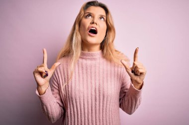Genç, güzel, pembe süveter giyen sarışın bir kadın izole edilmiş arka planda şaşkın şaşkın yukarı bakıyor ve parmaklarıyla işaret ediyor ve kollarını kaldırıyordu..