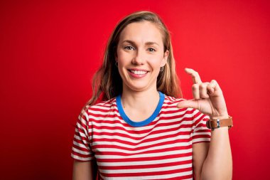 Young beautiful blonde woman wearing casual striped t-shirt over isolated red background smiling and confident gesturing with hand doing small size sign with fingers looking and the camera. Measure concept.