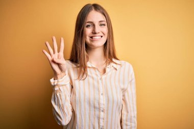 Genç, güzel, kızıl saçlı bir kadın, sarı arka planda günlük çizgili tişört giyiyor. Dört numarayı işaret ediyor ve kendinden emin ve mutlu bir şekilde gülümsüyor..