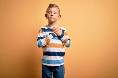 Mavi gözlü, sarı arka planda renkli çizgili gömlek giyen, göğsünde elleri kapalı, yüzünde minnet dolu bir jest olan beyaz bir çocuk. Sağlık kavramı.