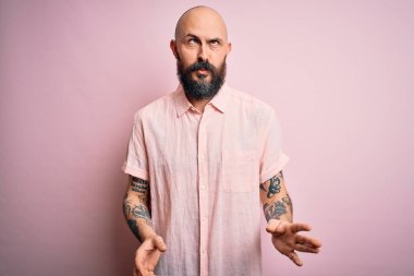 Sakallı ve dövmeli yakışıklı, pembe arka planda günlük gömlekler giyen, dudaklı, garip ve komik yüzlü bir adam. Komik ifade.