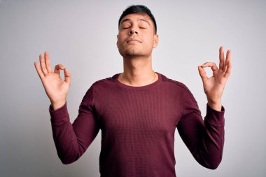 Beyaz, izole edilmiş arka planda dikilen gündelik tişört giyen genç, yakışıklı İspanyol adam rahatla ve gözleri kapalı, parmaklarıyla meditasyon hareketi yaparken gülümse. Yoga konsepti.