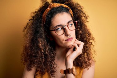 Güzel esmer kadın tatilde, kıvırcık saçlı ve piercing takıyor, gözlük takıyor ve eli çenesinde soru soran düşünceli bir ifade takınıyor. Düşünceli bir yüzle gülümsüyor. Şüphe kavramı.